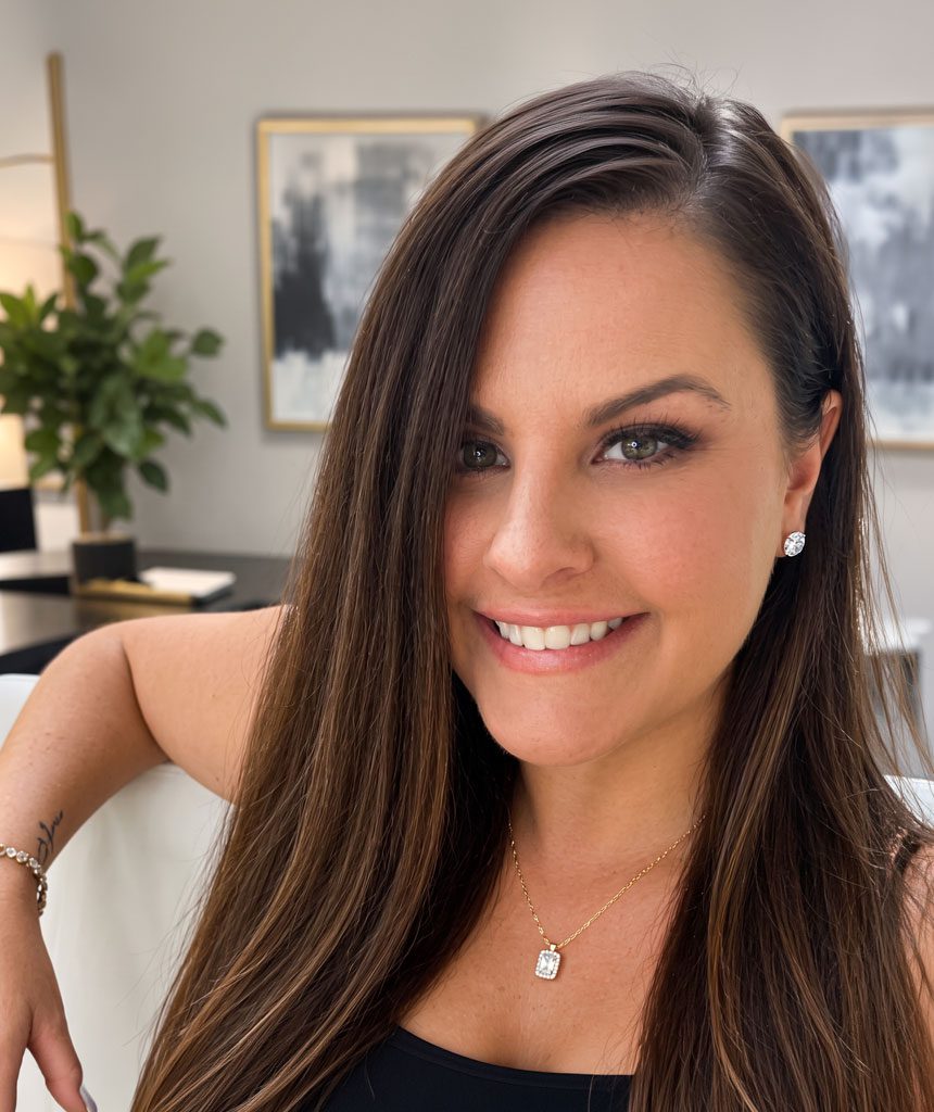 Smiling woman with long brown hair wearing diamond stud earrings and a gold pendant necklace in a well-lit indoor setting.