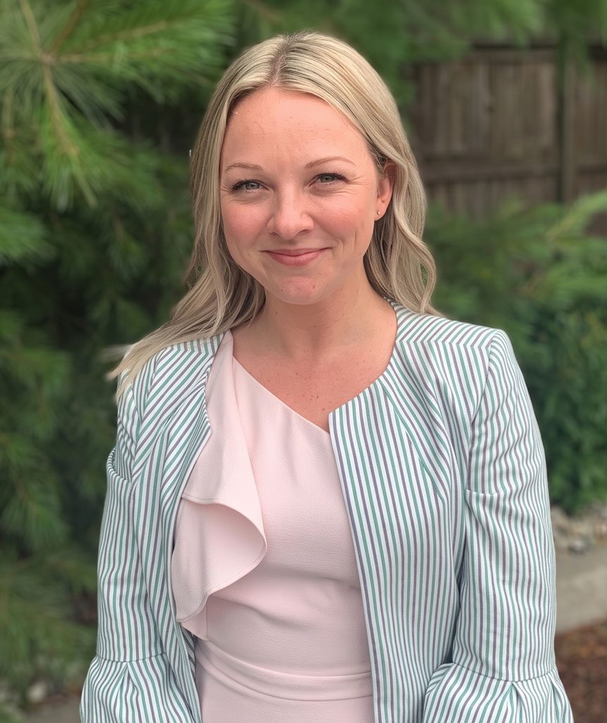 Portrait of a smiling blonde woman outdoors, wearing a pale pink blouse and a striped blazer with greenery in the background.