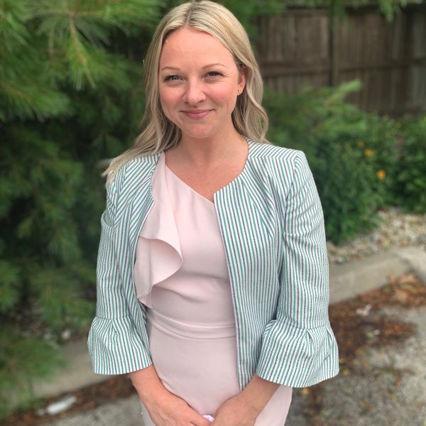 Smiling woman in a pale pink dress and green striped blazer, standing outdoors.